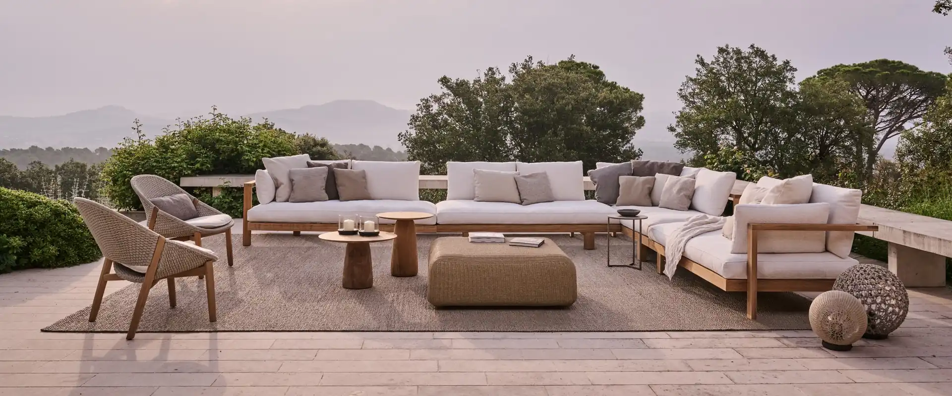 Tribù Pure teak corner sofa with white cushions, lounge chairs and a pouf on a terrace above wooded hills