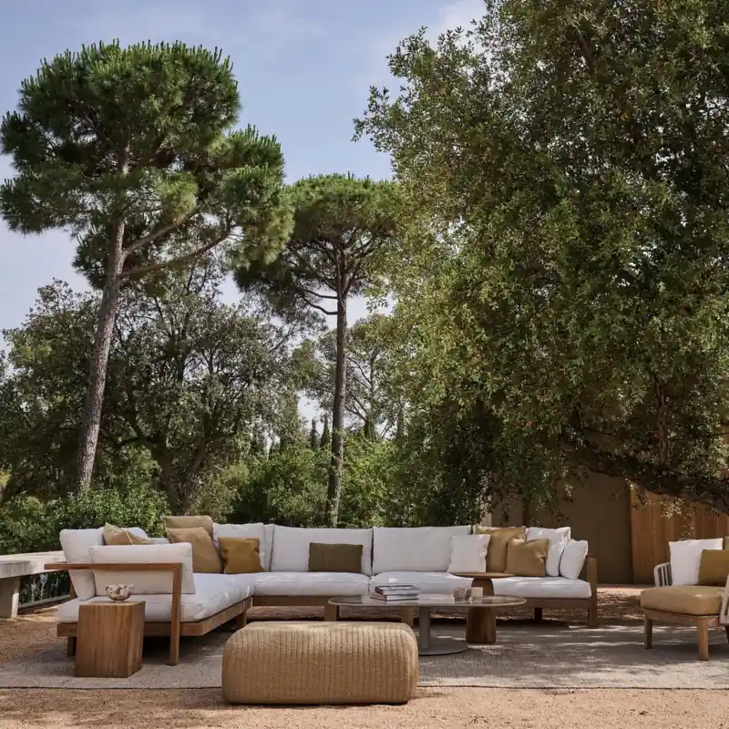 Tribù Pure teak sofa with white cushions, a woven pouf and an armchair on a terrace shaded by pine trees