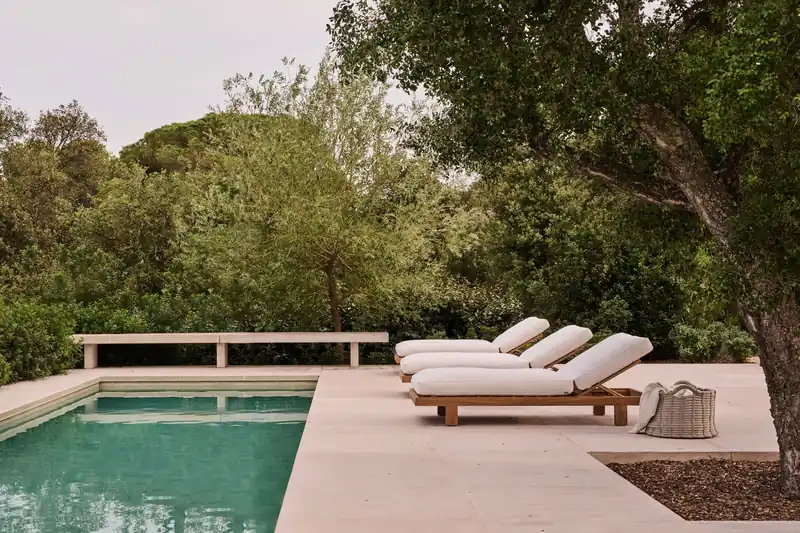 Three Tribù Pure sun loungers with white cushions on a stone deck beside a green pool, with pine trees behind