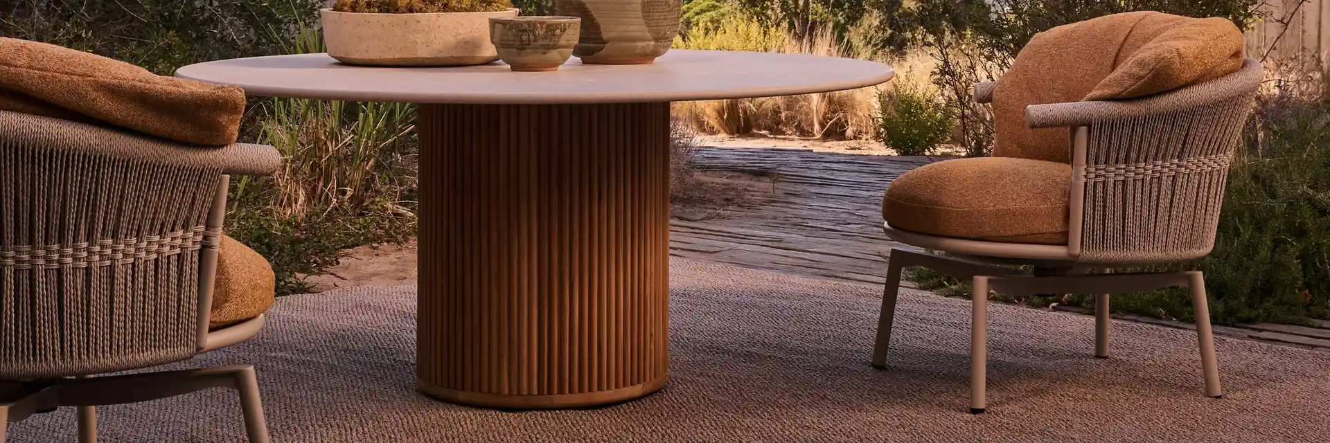 Tribù round dining table with a fluted teak base and rope-woven armchairs on a terrace in low sun
