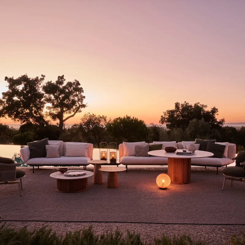 Tribù Senja sofas and lounge chairs around round tables with lanterns on a terrace at dusk, trees against a pink sky