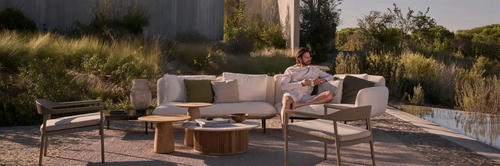 Man sitting on a Tribù Senja sofa in a garden of grasses and pines beside a concrete house