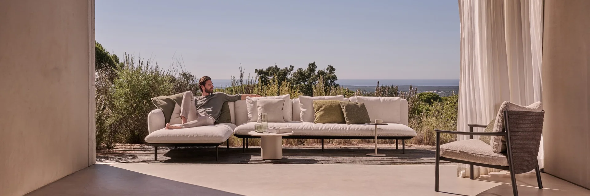Man lying on a Tribù Senja sofa on a terrace seen from under a pitched roof, with the sea on the horizon