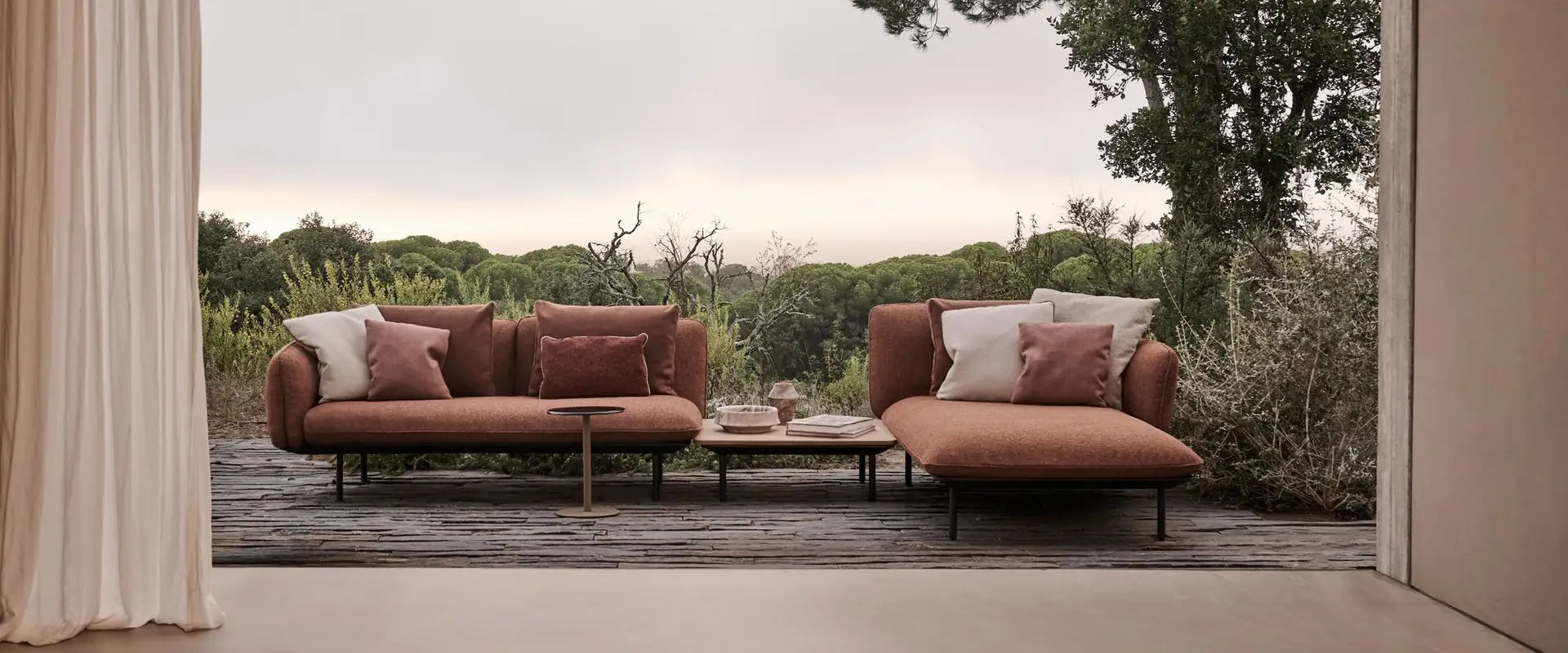 Two Tribù Senja sofas in terracotta fabric under a gabled porch with linen curtains, an umbrella pine outside