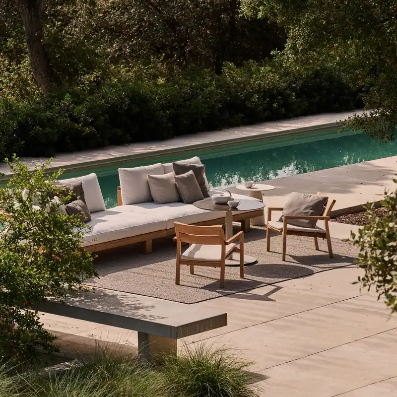 Tribù teak sofa and lounge chairs on a stone terrace beside a turquoise pool, surrounded by lush planting