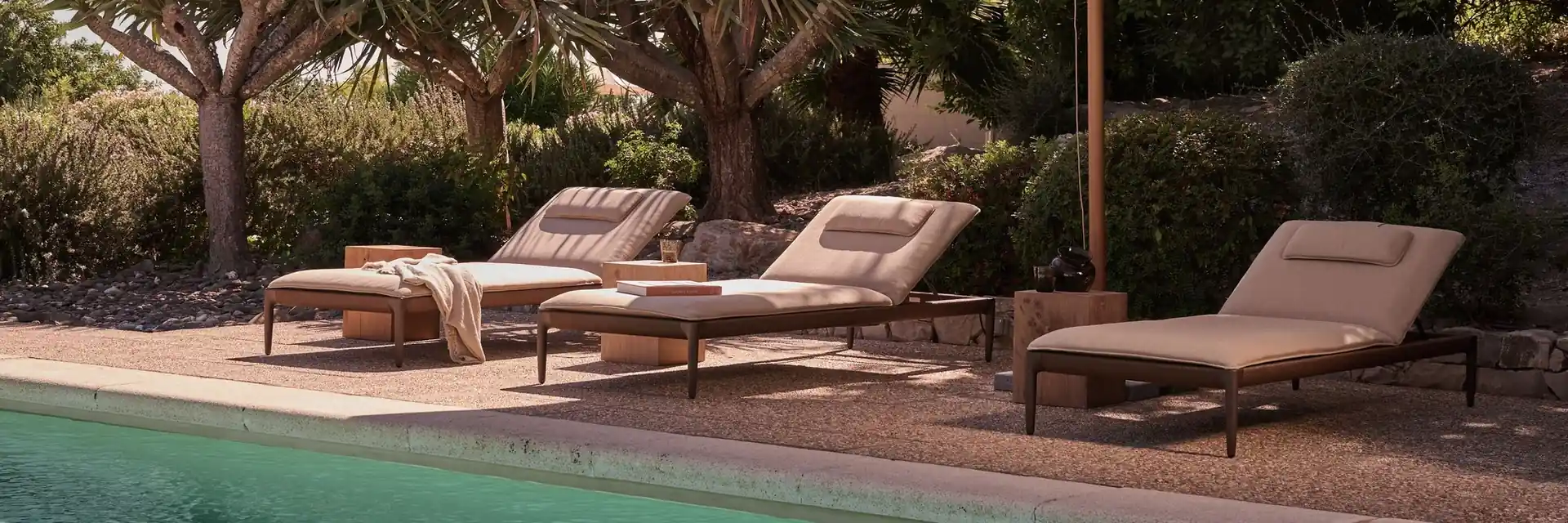 Tribù teak sun loungers with beige mattresses under a parasol beside a green pool, with dragon trees behind