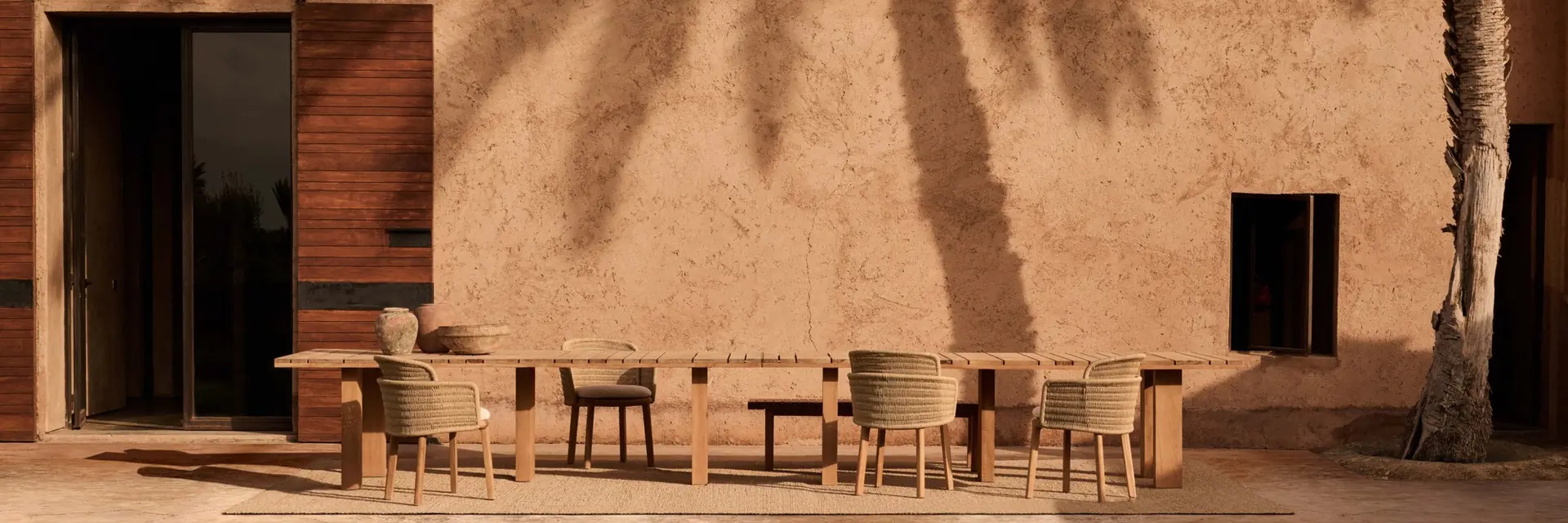 Tribù Suro long dining table and woven chairs in front of an adobe house, with the shadow of a palm tree on the wall