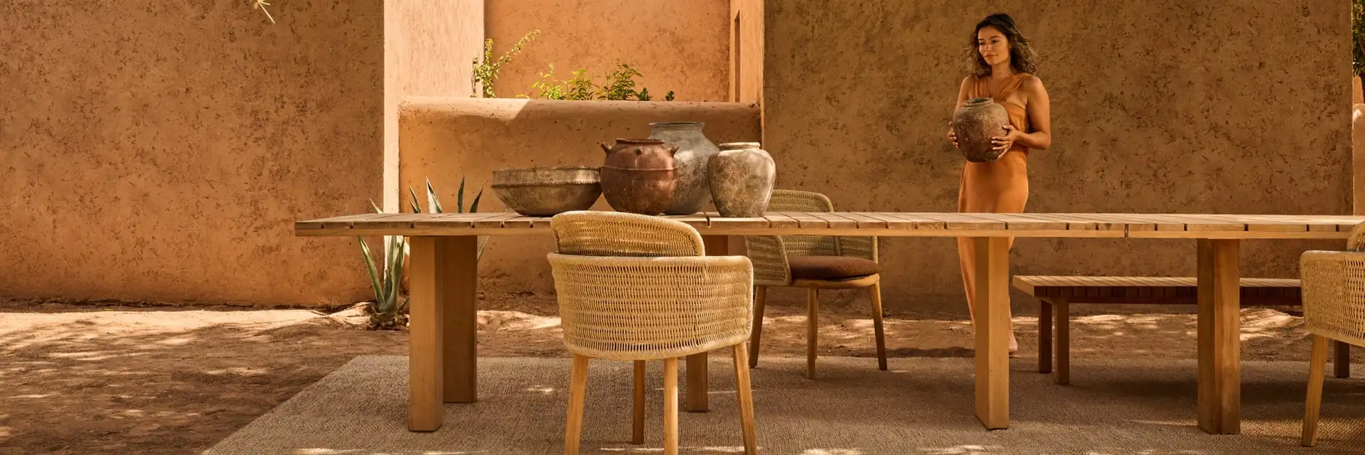 Woman standing at a long Tribù Suro dining table with Suro armchairs under a tree, beside an ochre wall