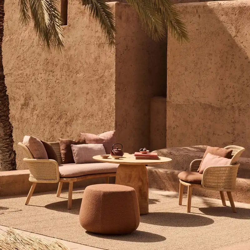 Tribù Suro woven lounge chairs and a sofa around a round table in a courtyard with earthen walls and a palm