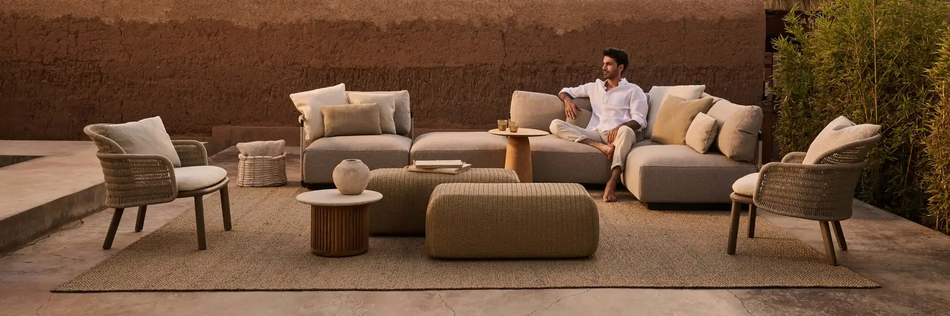 Man relaxing on a Tribù Suro sofa with armchairs and poufs beside an earth-coloured wall, with palm trees behind