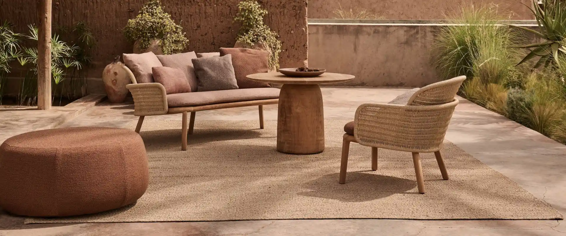Tribù Suro sofa, armchair and round table with a terracotta pouf on a terrace enclosed by earthen walls and plants