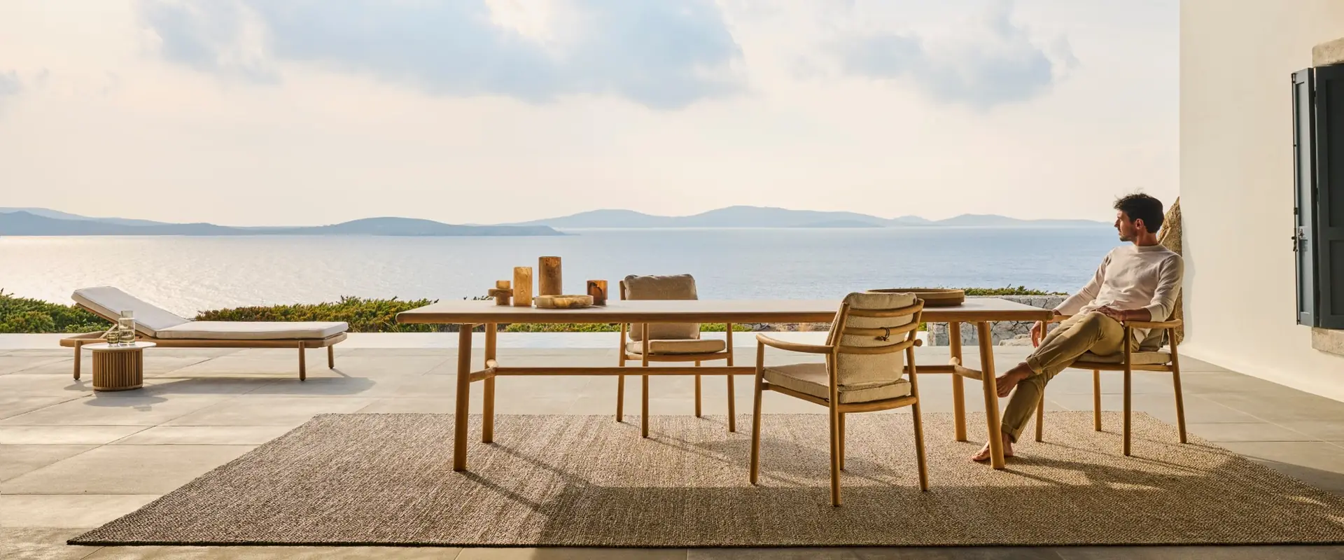 Man sitting at a Tribù teak dining table with woven chairs on a covered terrace, looking out over the sea and distant islands