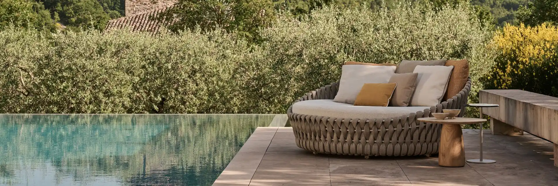Tribù Tosca round woven daybed with cushions beside an infinity pool, with olive trees and wooded hills behind