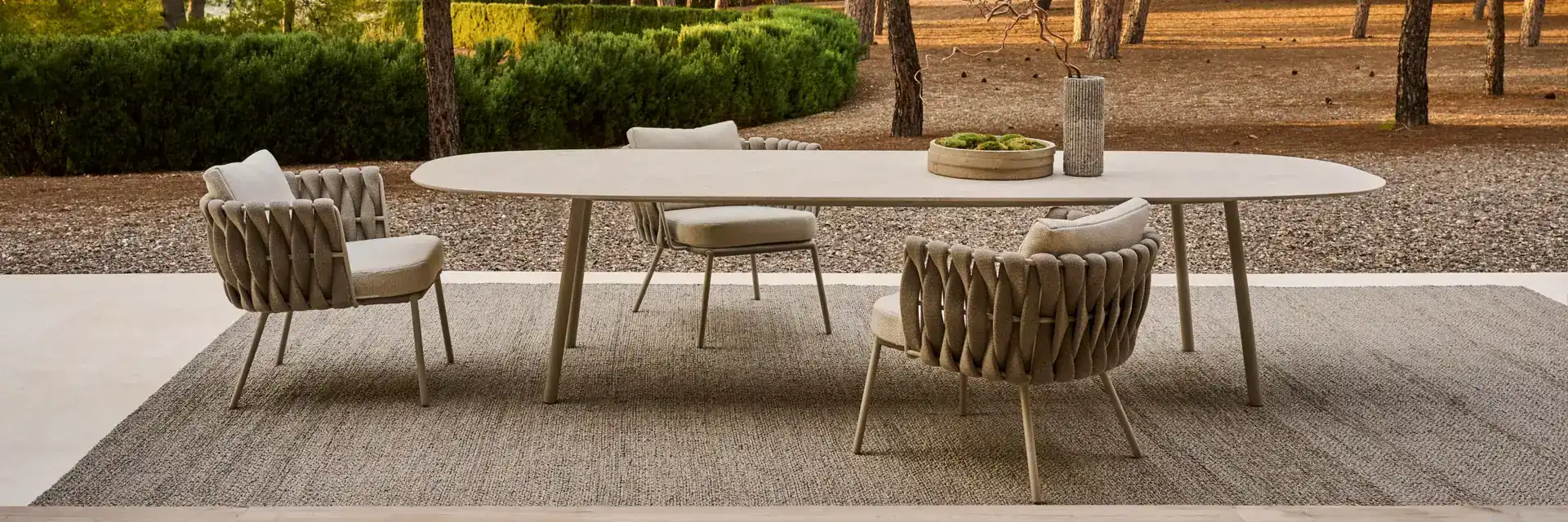 Tribù Tosca dining table and woven chairs on a rug in a garden of pine trees