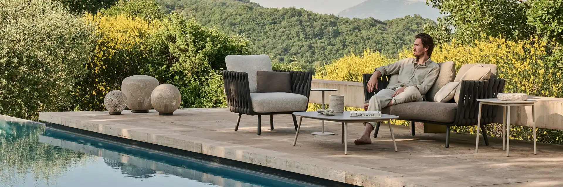 Man relaxing on a Tribù Tosca sofa beside an armchair and stone poufs on a pool terrace, with wooded hills behind