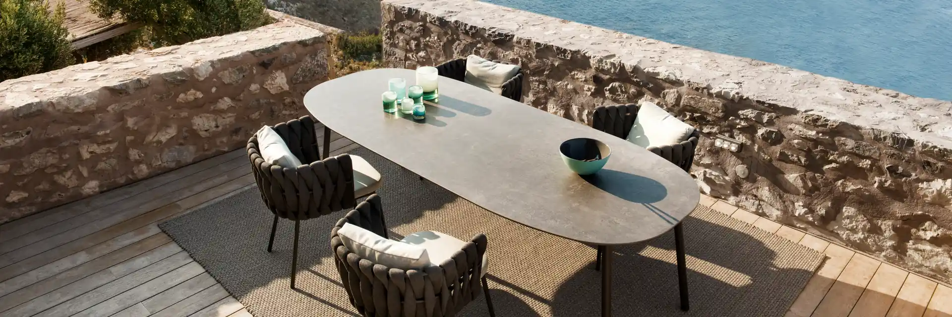 Tribù Tosca oval dining table and woven chairs on a stone terrace high above the sea and an island