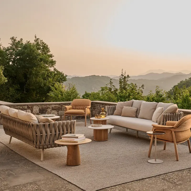 Tribù Tosca woven sofas and armchairs with round side tables on a terrace with a stone wall, hills at sunset