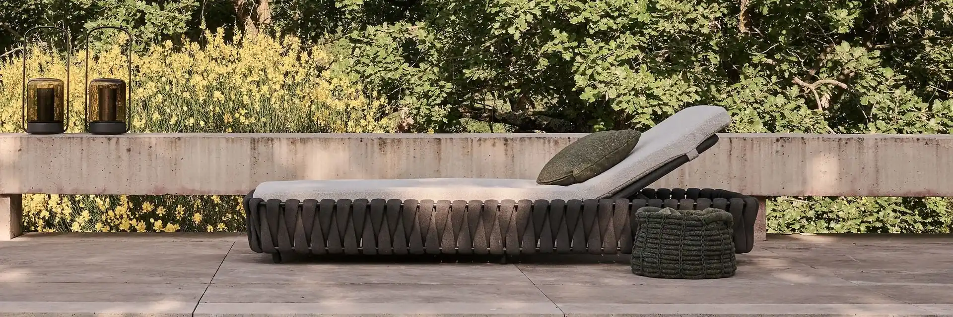 Tribù Tosca woven sun lounger on a stone platform beside a dark pool, with a green hedge behind