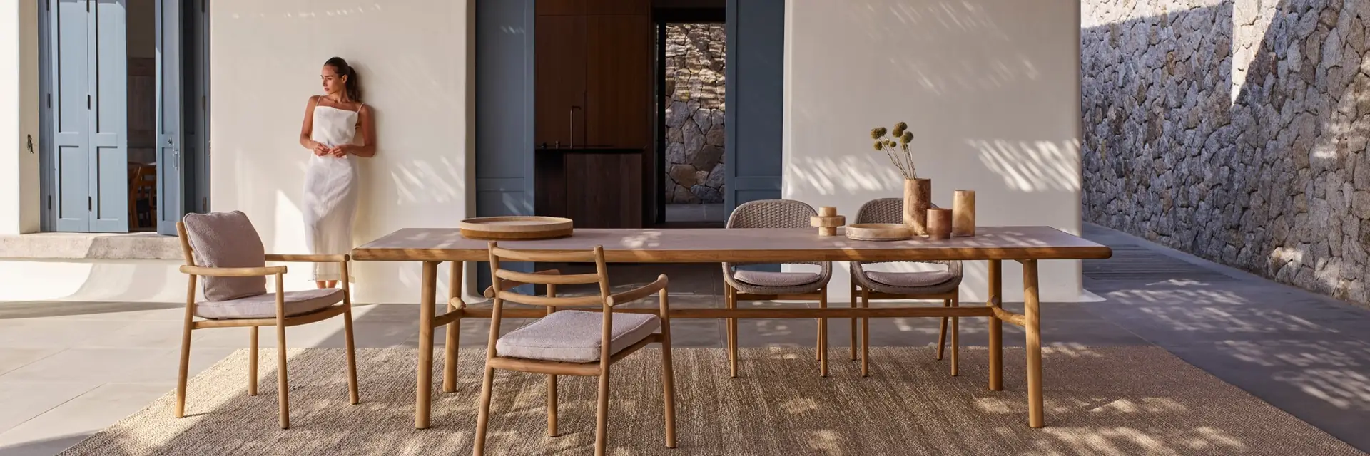 Tribù Ukiyo teak dining table and chairs on a terrace outside a white house with blue shutters, with a woman leaning on the wall