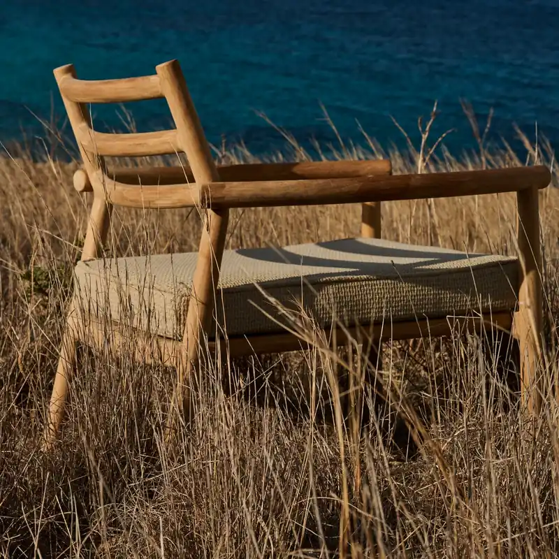 Tribù Ukiyo teak lounge chair standing in tall dry grass above a deep blue sea