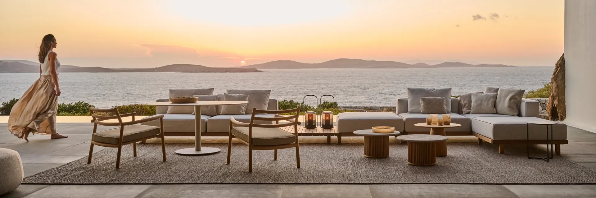 Tribù Ukiyo sofas and low tables under a timber pergola facing the sea at sunset, with a woman walking past