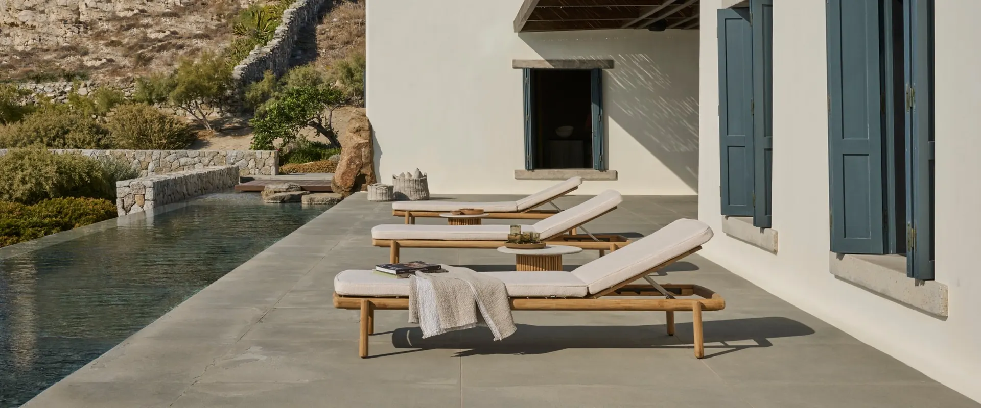 Tribù Ukiyo teak sun loungers along a narrow pool beside a whitewashed house with blue shutters and a rocky hillside