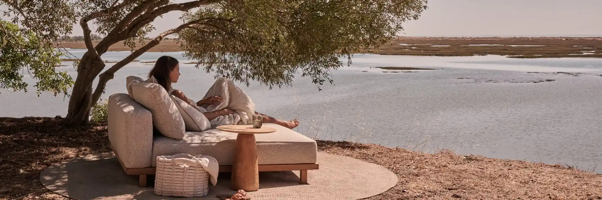 Woman sitting on a Tribù Vis-à-vis daybed under a tree beside a calm lagoon