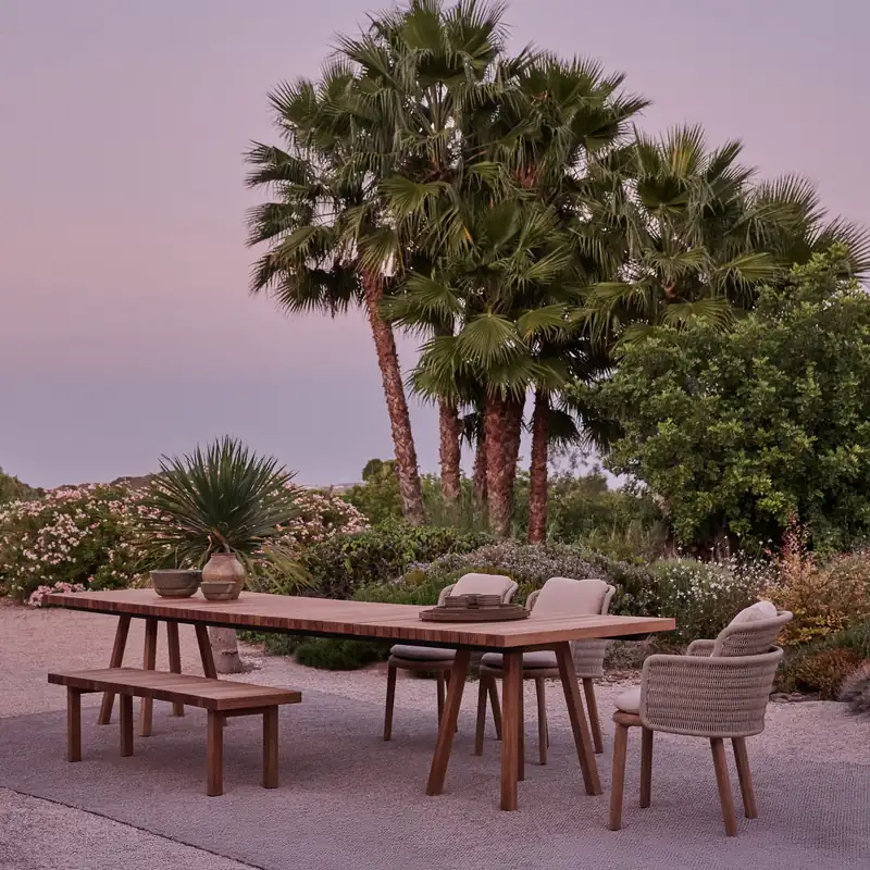Tribù Vis-à-vis teak dining table with a bench and woven chairs on gravel, with fan palms against a violet sky