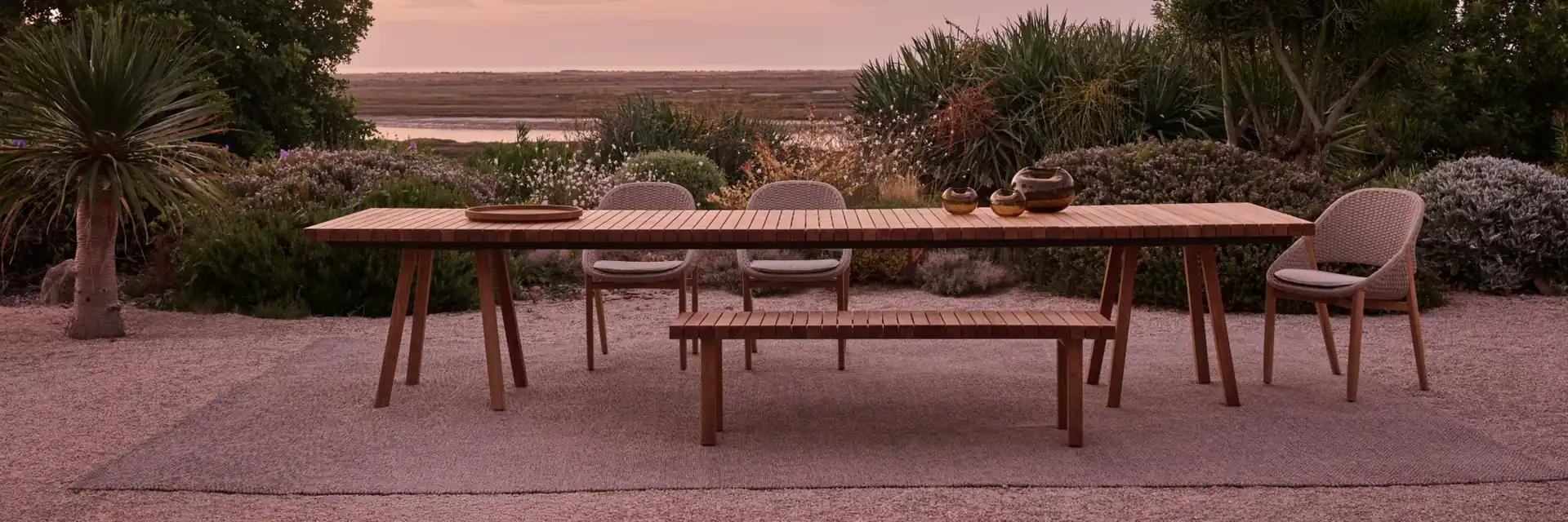 Tribù Vis-à-vis teak dining table with a bench and woven chairs on gravel, among shrubs under a pink sunset sky