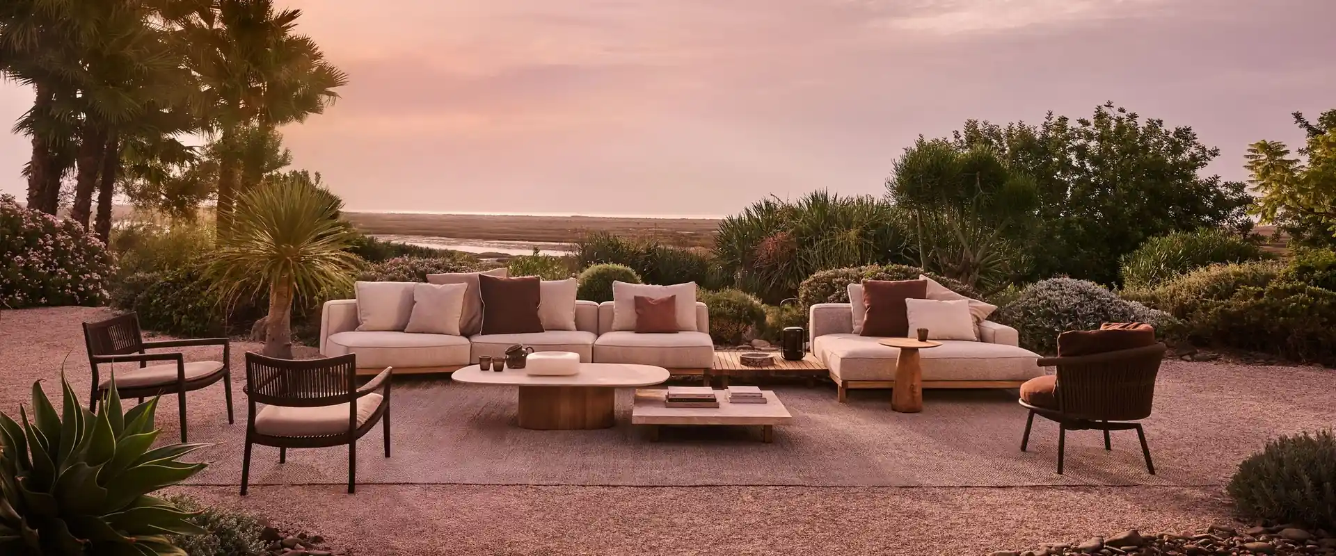 Tribù Vis-à-vis sofas and lounge chairs around low tables on gravel, among shrubs under a pink sunset sky