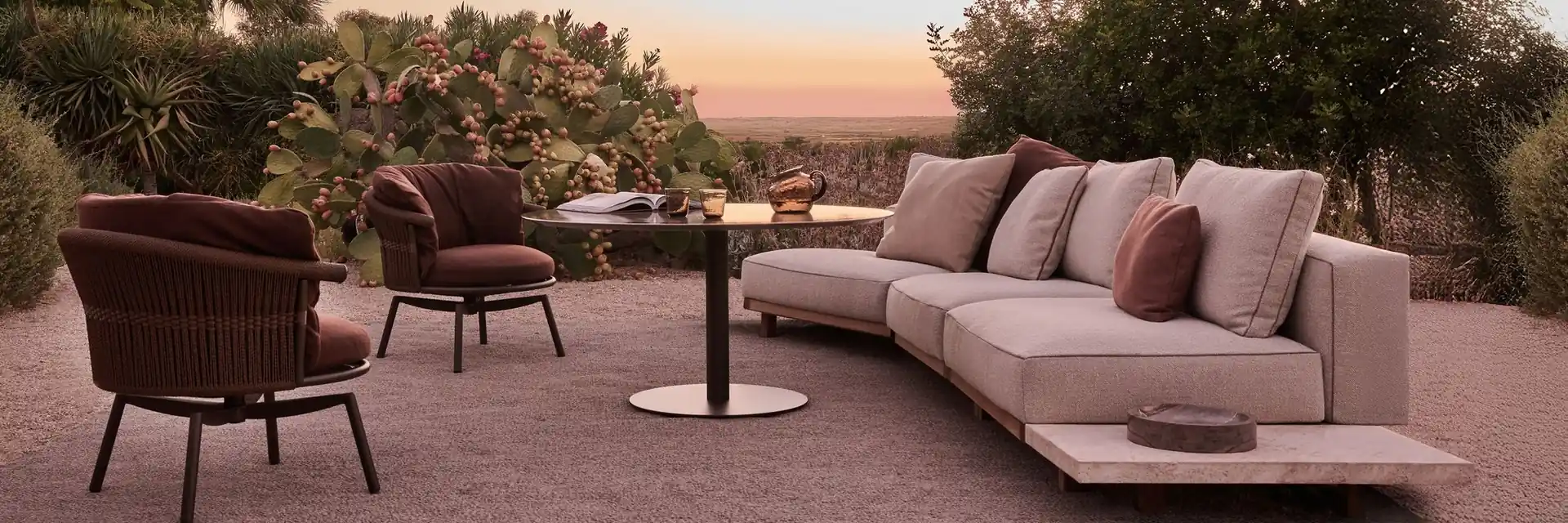 Tribù Vis-à-vis modular sofa and dark armchairs on a terrace at sunset, with trees behind