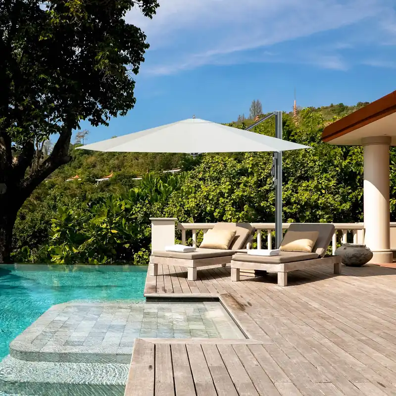 White Tuuci Bay Master M1 Classic Cantilever parasol shading two cushioned sun loungers on a wooden deck beside a turquoise pool, with trees behind