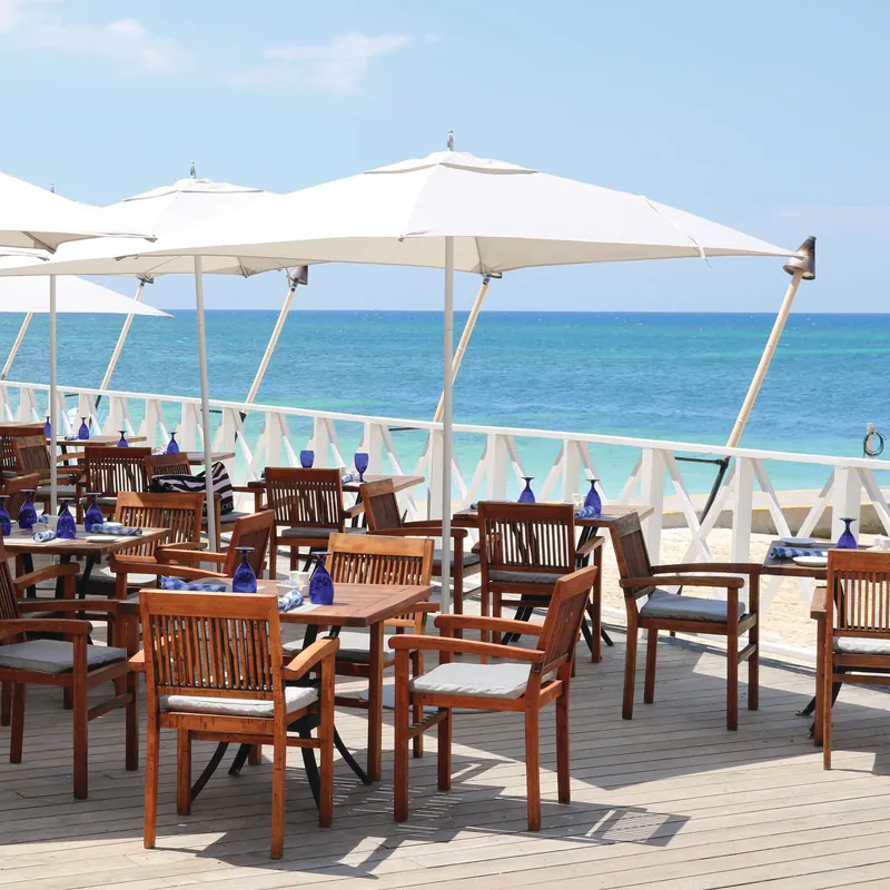 White square Tuuci Bay Master M1 Classic center post parasols shading wooden dining tables and chairs on a restaurant deck beside a turquoise sea