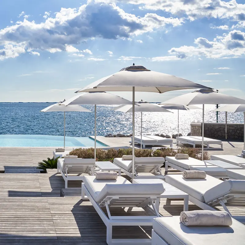 Tuuci Bay Master M1 Fiber-Flex center post parasols with pale canopies over white sun loungers on a wooden deck beside an infinity pool, with the sea and a cloudy blue sky behind