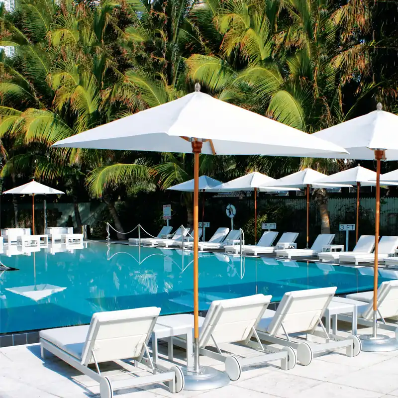 White Tuuci Bay Master M1 Maritime Hardwood center post parasols with hardwood poles shading rows of white sun loungers around a swimming pool, backed by tall palms