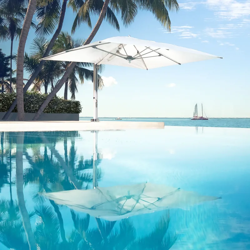 White Tuuci Bay Master M1 Swim Under Cantilever parasol opened over the water of an infinity pool and reflected in it, with palm trees, a sailboat and the open sea behind