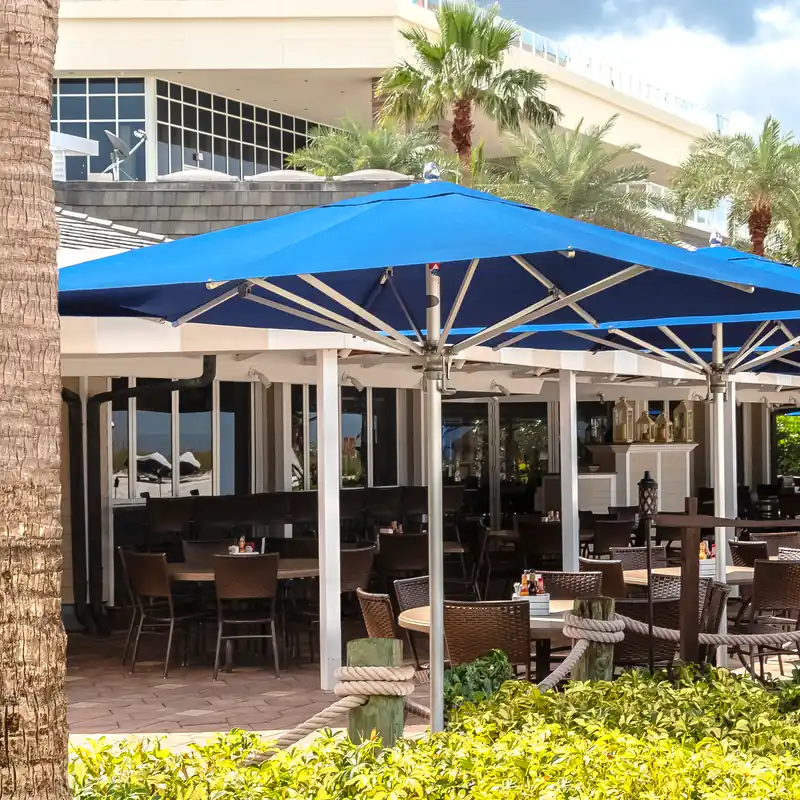 Large blue Tuuci Bay Master MAX Classic center post parasol with a satin anodized frame covering the tables of a restaurant terrace, with palm trees and a low hedge in front