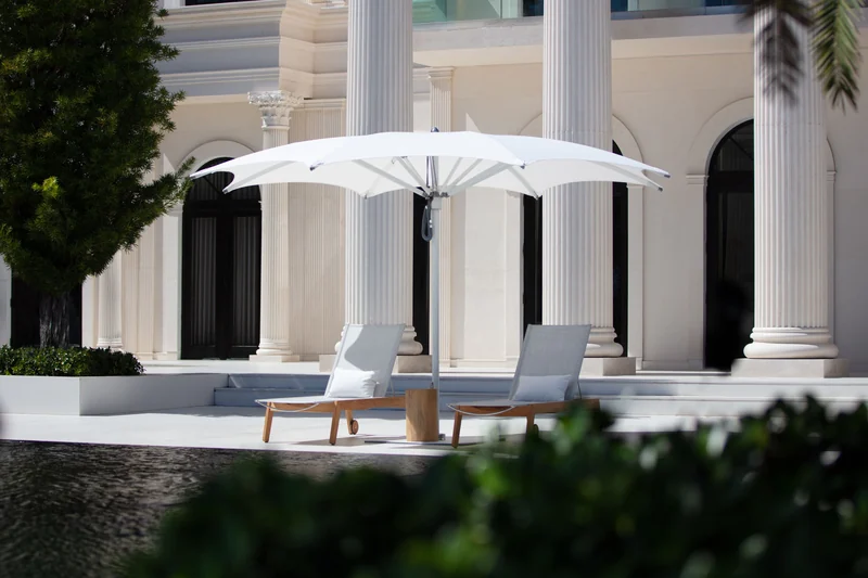 White Tuuci Ocean Master MAX Crescent parasol with a scalloped canopy over two loungers in front of classical columns