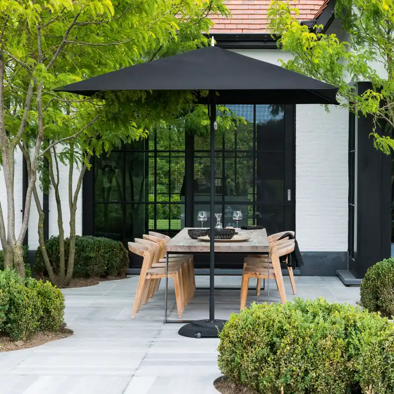 Black square Tuuci Ocean Master M1 Classic center post parasol over a wooden dining table and chairs on a stone terrace, with clipped topiary spheres and a white house behind