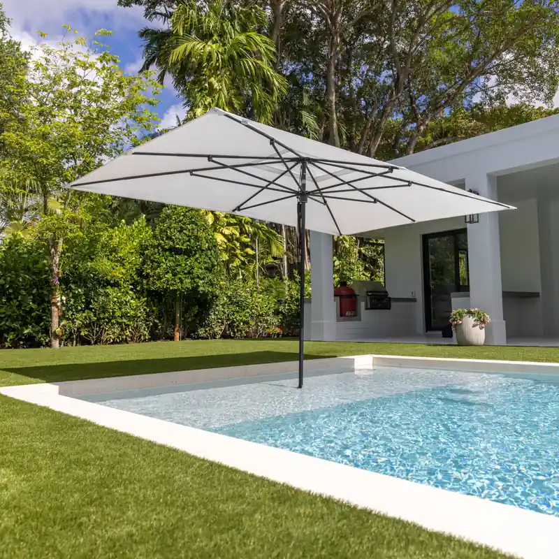 White Tuuci Ocean Master M1 Classic center post parasol with a graphite frame standing in the shallow end of a small swimming pool, in front of a white modern house with a lawn and trees
