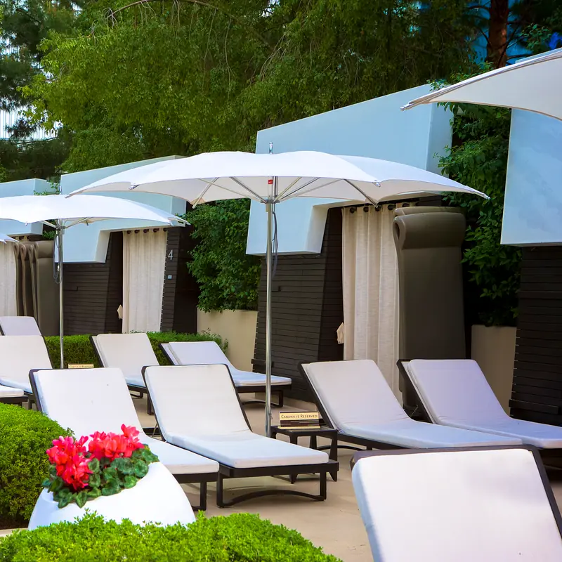 White Tuuci Ocean Master M1 Crescent center post parasols with curved canopies and polished masts shading rows of white sun loungers in front of curtained cabanas, with clipped hedges and red flowers