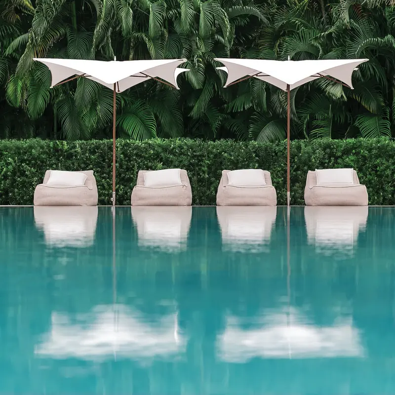 Two white Tuuci Ocean Master M1 Manta center post parasols over four cushioned lounge chairs at the edge of a pool, reflected in the water in front of a clipped hedge and palms