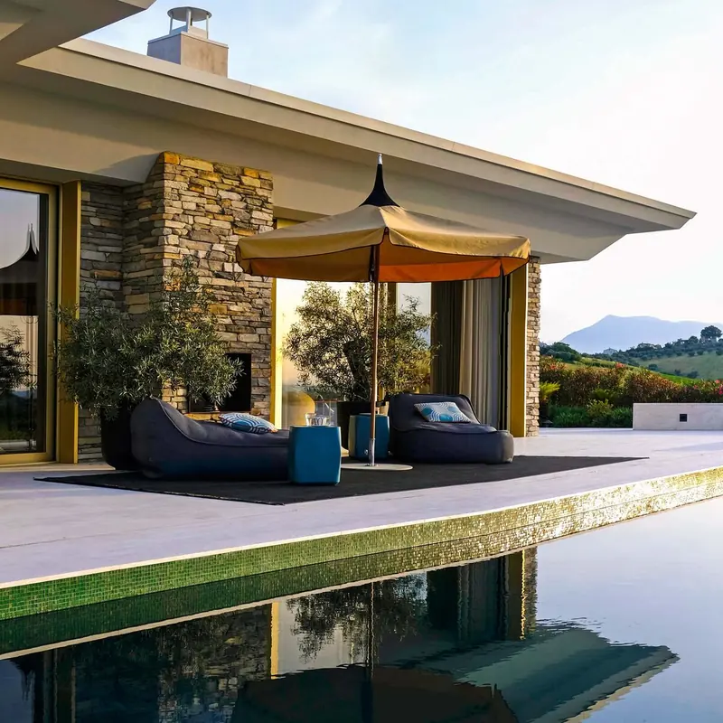 Tuuci Ocean Master M1 Pagoda center post parasol with a tan canopy and contrasting valance over lounge seating on the terrace of a stone villa, beside an infinity pool with rolling hills beyond