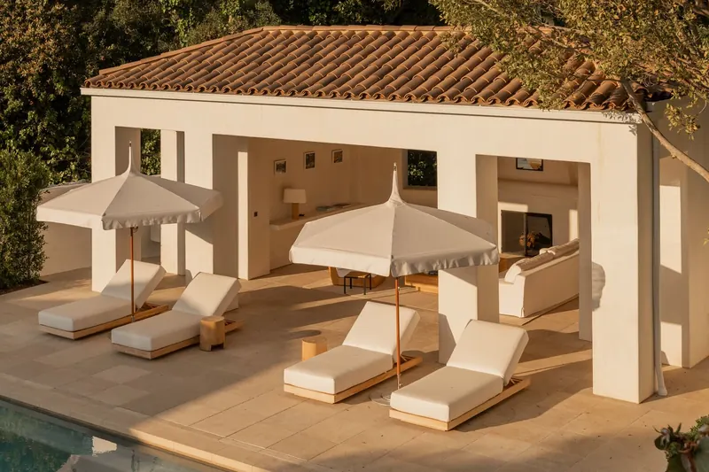 Two cream Tuuci Ocean Master M1 Pagoda parasols over cushioned sun loungers beside a white pool house with a terracotta tile roof, in warm evening light