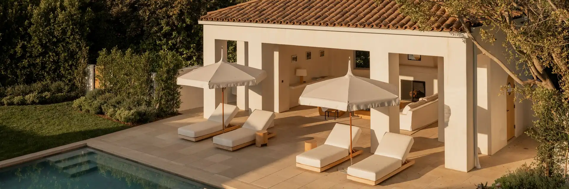 Two cream Tuuci Ocean Master M1 Pagoda parasols over cushioned sun loungers beside a white pool house with a terracotta tile roof, in warm evening light