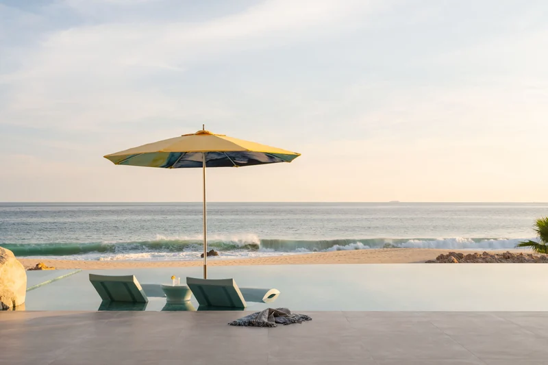 Yellow Tuuci parasol shading two in-pool loungers at the edge of an infinity pool, with waves breaking on the beach beyond at sunset
