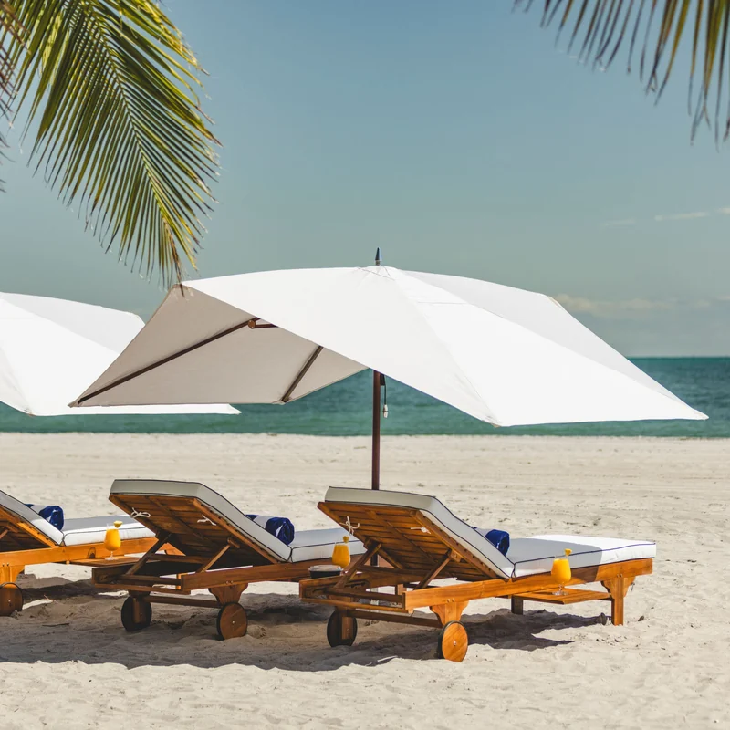 White Tuuci Ocean Master M1 Polynesian center post parasol with a peaked canopy over teak sun loungers on a sandy beach, with palm fronds overhead and the sea behind