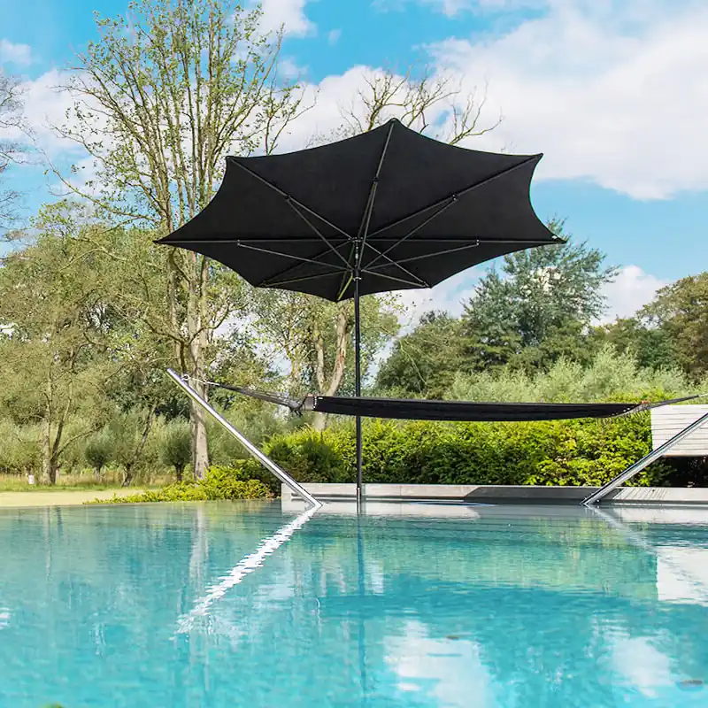 Black Tuuci Ocean Master M1 Razor center post parasol with a pointed, asymmetrical canopy standing in a swimming pool beside a hammock lounge, with a lawn and trees behind
