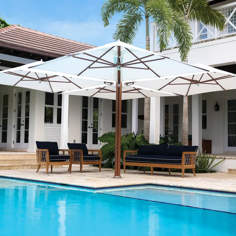 Tuuci Ocean Master M1 Shade Pod Cantilever parasol with four white canopies on a single wood-finish mast shading teak seating beside a swimming pool, in front of a white house with a veranda