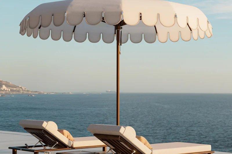 Tuuci Ocean Master M1 Voilare center post parasol with a scalloped cream canopy and pointed top over two cushioned sun loungers on a terrace above the sea